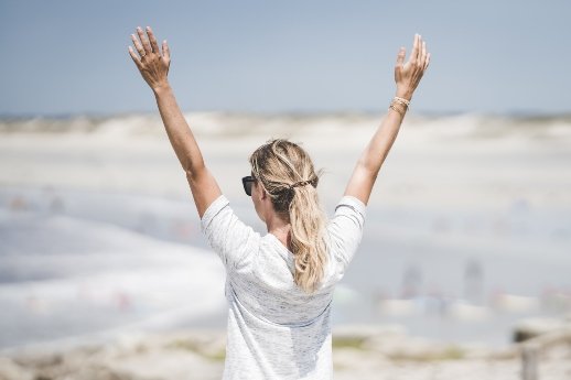 WOOP-Methode . Frau in weißem Hemd am Strand hebt die Arme in die Luft