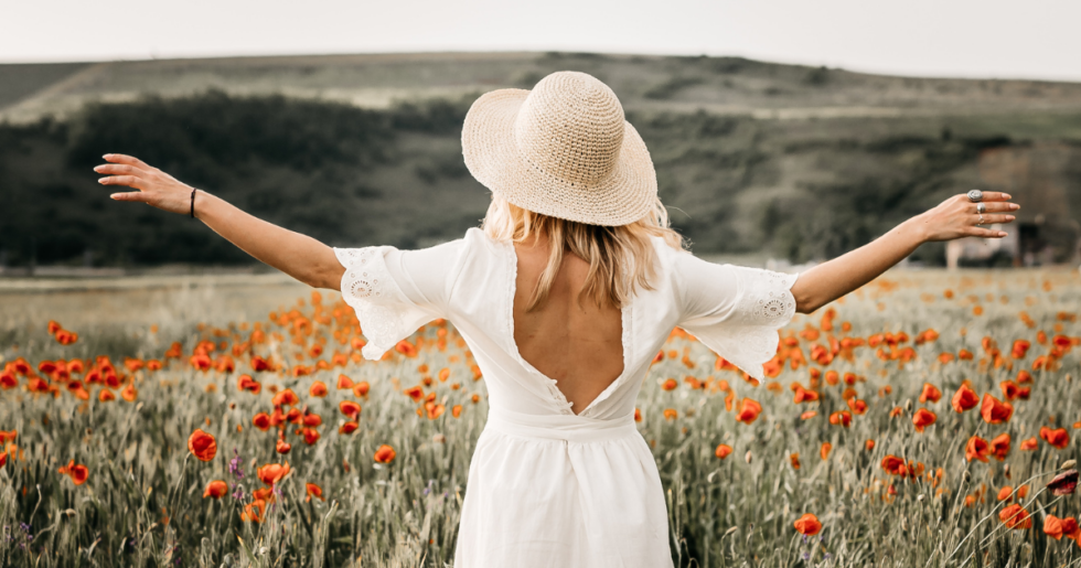 Frau in weißem Kleid und Strohhut breitet die Arme in einem Mohnblumenfeld aus, symbolisiert Freiheit und Loslassen.