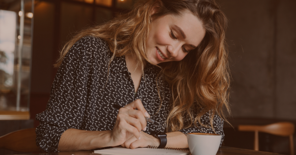 Frau sitzt lächelnd in einem Café und schreibt in ihr Notizbuch, während sie eine Tasse Kaffee genießt – so gut kann sich Braindumping anfühlen.