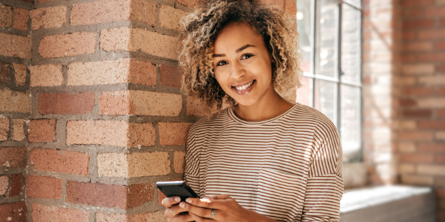 Lächelnde Frau mit Smartphone, steht an einer Backsteinmauer, symbolisiert positive Montagmorgen-Impulse durch den Newsletter von Freedom Lover.