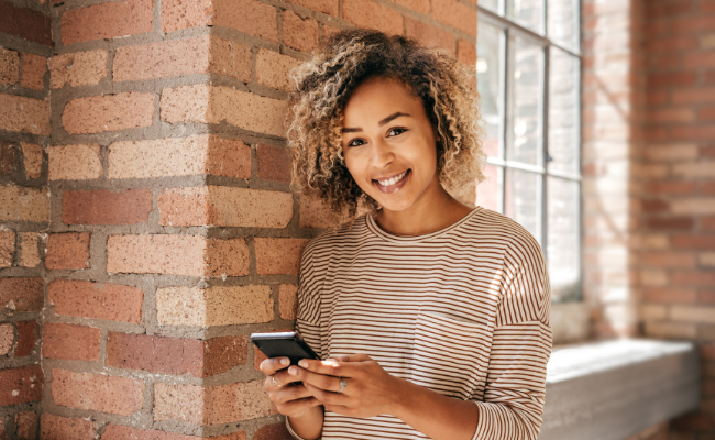 Lächelnde Frau mit Smartphone, steht an einer Backsteinmauer, symbolisiert positive Montagmorgen-Impulse durch den Newsletter von Freedom Lover.