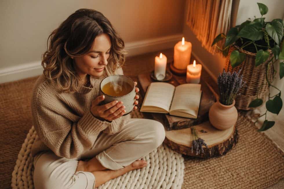 Frau sitzt abends mit einer Tasse Tee auf einem Bodenkissen neben Kerzen und einem offenen Buch.