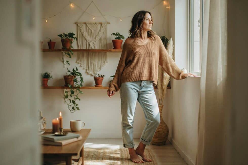 Frau im Boho Style steht am Fenster und sieht nachdenklich raus
